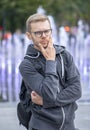 Portrait of a thoughtful man in glasses 25-30 years old with a backpack on his shoulder against a blurry background of city founta Royalty Free Stock Photo
