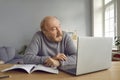 Portrait of thoughtful red-haired elderly man with beard, holding glasses and sitting at desk at home with laptop and Royalty Free Stock Photo
