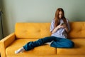 Portrait of thoughtful redhead young woman looking at smartphone screen sitting on yellow sofa, chatting online, using Royalty Free Stock Photo