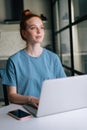 Portrait of thoughtful redhead young woman working typing on laptop computer sitting at table Royalty Free Stock Photo