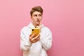 Portrait of thoughtful student boy in white hoodie on pink background holding smartphone, looking away at copy space with pensive Royalty Free Stock Photo