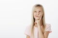 Portrait of thougtful creative young girl with long blond hair, looking up and holding hand on chin while thinking or