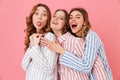 Portrait of three beautiful young girls 20s wearing colorful striped pyjamas having fun at slumber party, isolated over pink back