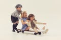 Portrait of three cheerful children playing together, boy rolling girls on skateboard isolated over grey studio Royalty Free Stock Photo