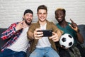 Portrait of three happy young men taking a selfie while sitting at home with beer and snacks on sofa watch tv Royalty Free Stock Photo