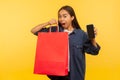 Portrait of thrilled amazed shopper girl in denim shirt holding bags and mobile phone with empty display mock up