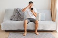 Portrait of tired brunette man wearing white t shirt and jeans shirt sitting on sofa at home, holding mobile phone and looking at Royalty Free Stock Photo