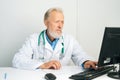 Portrait of tired mature adult male doctor wearing white coat with stethoscope working on desktop computer sitting at Royalty Free Stock Photo