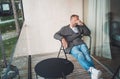 Portrait of a tired Middle-aged man dressed in open cardigan, jeans, and warm socks sitting on forest house balcony. He massaging