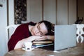 Portrait of tired middle-aged woman sitting at table near laptop, sleeping on crossed arms on pile of book sat home.