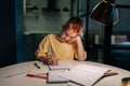 Portrait of tired schoolboy studying at home doing homework sitting at table under light of lamp at evening. Front view Royalty Free Stock Photo