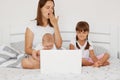 Portrait of tired sleepy woman mother sitting on bed with her children and working on laptop, yawning, covering mouth with palm, Royalty Free Stock Photo