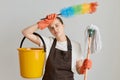 Portrait of tired woman wearing brown apron and white t shirt cleaning her flat, holding mop, ppduster and bucket, keeping hand on