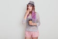 Portrait of tired young woman wearing hat, having bright violet hair, holding disposable cup with coffee, needs energy to wake up Royalty Free Stock Photo