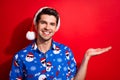 Portrait of toothy beaming person dressed santa hat blue shirt palm show object empty space isolated on vibrant red