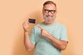 Portrait of toothy beaming senior man in glasses wear stylish t-shirt indicating at debit card isolated on pastel color