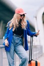 Portrait of a traveler woman standing with an orange suitcase near an airport. Young fashionable woman in a blue jeans