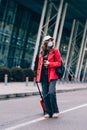 Portrait of a traveler woman in a mask walking with an orange suitcase near an airport. Young fashionable woman in a