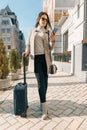 Portrait of traveling young woman with mobile phone and suitcase, fashionable girl on the city street, wearing warm coat, sunny Royalty Free Stock Photo