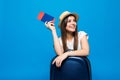 Portrait of trendy young female in wicker hat standing with suitcase and holding passport with tickets, over blue background Royalty Free Stock Photo