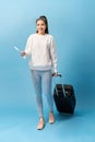 Portrait of trendy young girl standing with suitcase and holding passport with tickets, over blue background Royalty Free Stock Photo