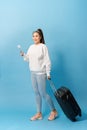 Portrait of trendy young girl standing with suitcase and holding passport with tickets, over blue background Royalty Free Stock Photo