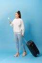 Portrait of trendy young girl standing with suitcase and holding passport with tickets, over blue background Royalty Free Stock Photo