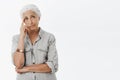 Portrait of troubled and concerned gloomy senior woman with white hair smirking holding finger on temple and looking at