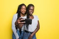 Portrait of two young african girls smiling and waving hand at camera while taking selfie on mobile phone isolated over yellow Royalty Free Stock Photo