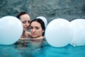 Portrait of two attractive young smiling women, girlfriends hiding wet among white balloons, while bathing in the spa pool Royalty Free Stock Photo