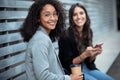 Two beautiful businesswomen using smart phone while looking at camera sitting on a bench on the street