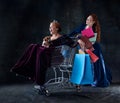 Portrait of two beautiful women in image of queens isolated over dark background. Cheerful royal shopping Royalty Free Stock Photo