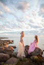 Portrait of two beautiful young gorgeous women wearing fashionable silk dress against stone, sunset sky and clouds