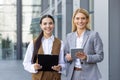 Portrait of two businesswomen standing outside the office center in business clothes, holding a tablet and a folder with Royalty Free Stock Photo