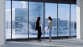 Portrait of two busineswoman in office standing in front of office buildings behind the window. Coworkers discussing