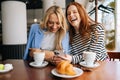 Portrait of two cheerful beautiful women friends sitting in cafe indoors, looking at phone and talking, laughing. Happy Royalty Free Stock Photo