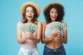 Portrait of two excited women with different type of skin wearing straw hats and summer clothing looking at lots of money holding Royalty Free Stock Photo