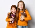 Portrait of two frolic girls friends in orange shirts standing together with dolls hugging sticking out their tongues Royalty Free Stock Photo