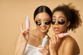 Portrait of two funny young women holding bottles with beauty product, posing with applied black hydro gel under eye