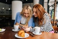 Portrait of two happy young women sitting at table with coffee at cafe by window, having conversation and showing each Royalty Free Stock Photo