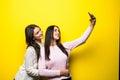 Portrait of two lovely girls dressed in sweaters standing and taking a selfie over yellow background Royalty Free Stock Photo