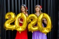 Portrait of two smiling happy girls in christmas dresses posing with gold colored number balloons 2020