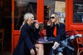 Portrait of two young beautiful women friends wearing black trench coats, sitting at table outside red cafe, talking. Royalty Free Stock Photo