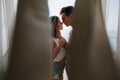 Portrait of two young people in love with white curtains on foreground. Man with dark hair holding his girlfriend`s face Royalty Free Stock Photo