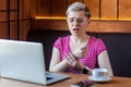 Portrait of unhappy young bussinesswoman with short blonde hair in pink t-shirt and eyeglasses sitting in cafe and holding arm