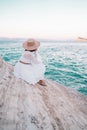 Portrait of unrecognizable woman in white boho dress, rope trendy sandals and straw hat sitting alone on clay beach Royalty Free Stock Photo