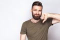 Portrait of unsatisfied bearded man with thumbs down and dark green t shirt against light gray background.