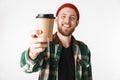 Portrait of unshaved man wearing hat and plaid shirt holding paper cup with coffee, while standing isolated over white background