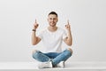 Portrait of upbeat good-looking man in trendy eyewear, sitting on floor with crossed legs, pointing up with both index Royalty Free Stock Photo