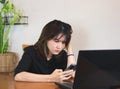 Upset Asian woman wearing black shirt, sitting at wooden table with computer laptop, reading message in mobile phone, touching Royalty Free Stock Photo